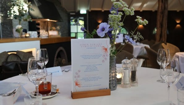 Gedekte tafel in een restaurant met bloemen, servies e.d. op tafel en een menukaart in een houten houder van het Diner Pensant.