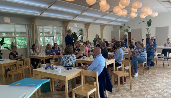 Zaal vol met mensen aan houten tafels, twee personen staan.
