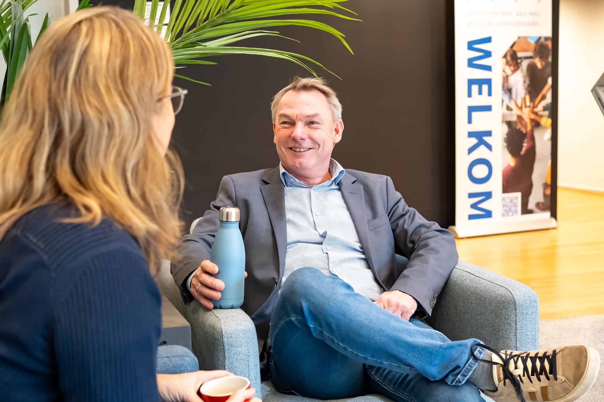 Je ziet een witte man die lachend in een stoel zit. Hij heeft een grijs jasje, een lichtblauw overhemd en een spijkerbroek aan. Links kijk je op de rug van een vrouw met lang, donkerblond haar, een bril en een donkerblauwe trui. Op de achtergrond zie je een donkergrijze muur en een banner met 'WELKOM' in blauwe letters.