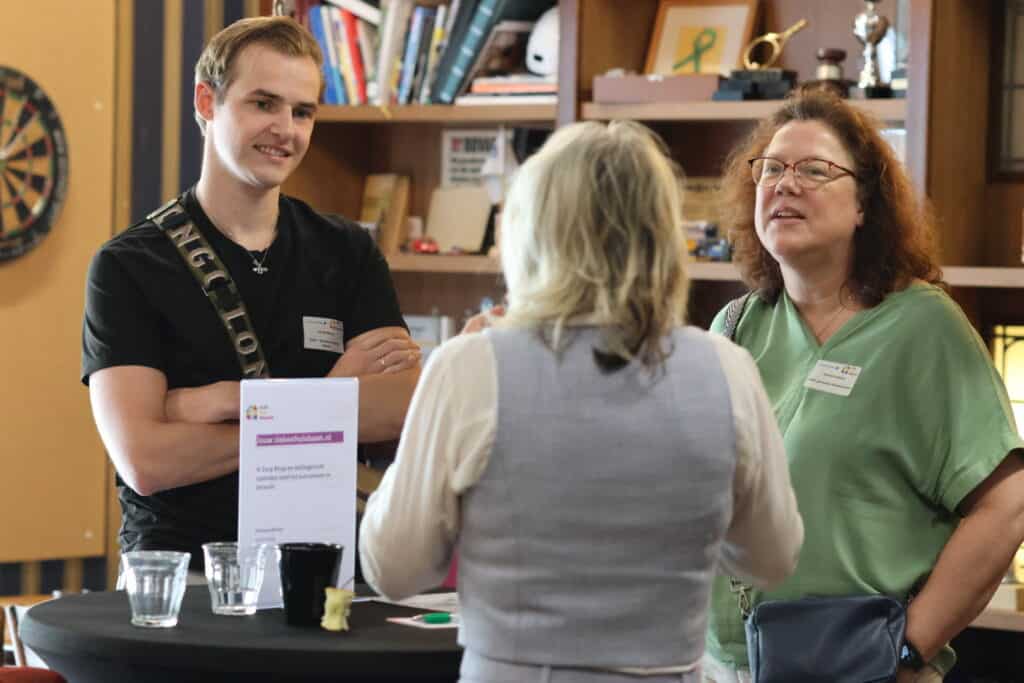 Drie personen staan rondom een tafel te kletsen. Twee kijk je in het gezicht, de ander op de rug.