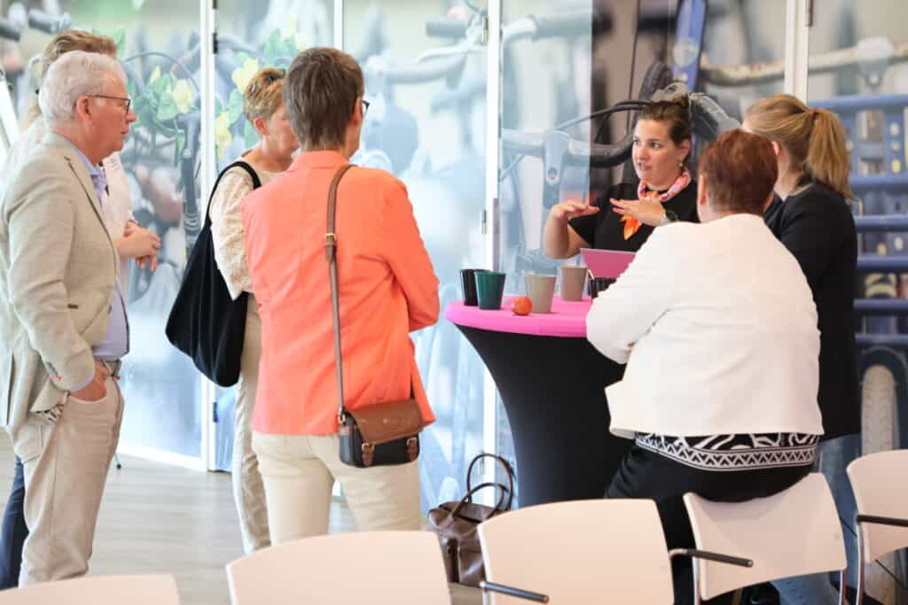 Een groep mensen staat om een statafel heen. Vijf anderen luistern naar hoe een vrouw geanimeerd vertelt.