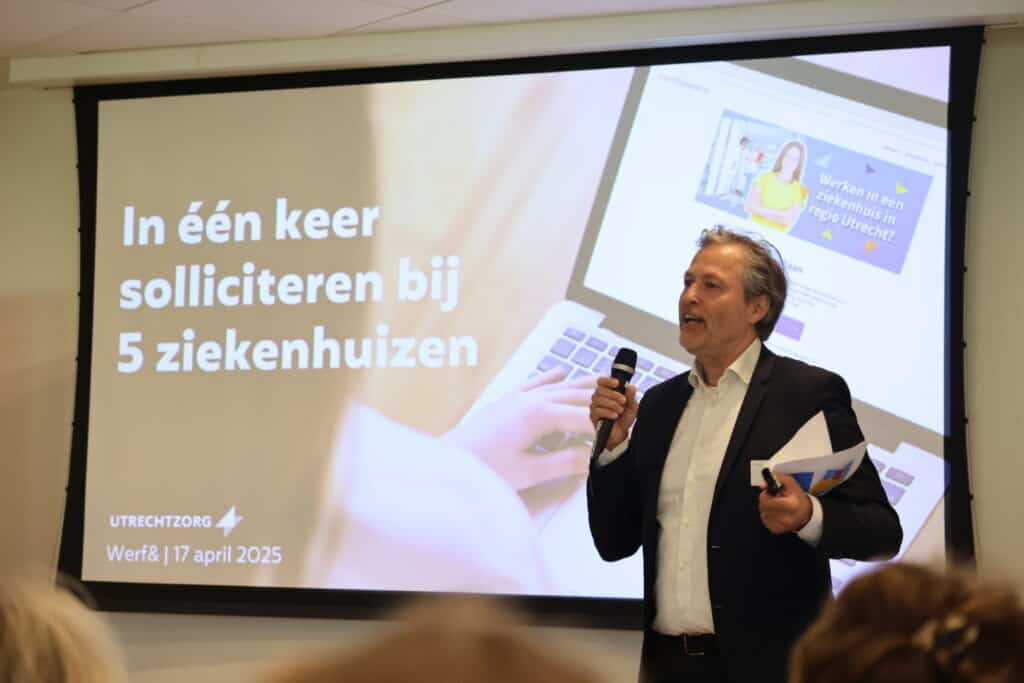 Een man staat voor een groot scherm.  Hij houdt een microfoon in zijn handt en is iets aan het vertellen. Hij heeft een pak aan. Op het scherm staat "In één keer solliciteren bij 5 ziekenhuizen".