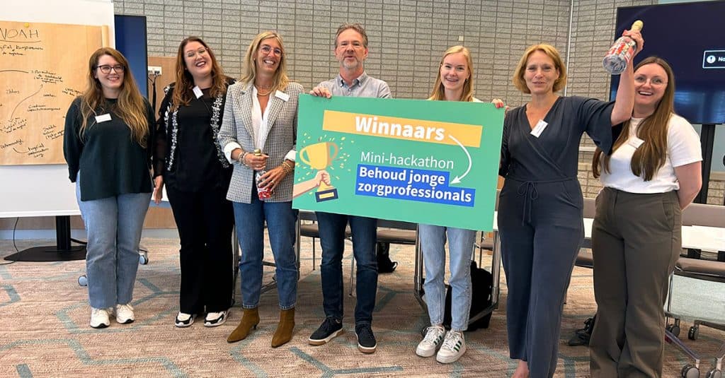 7 personen staan op een rijtje. Twee houden een bord vast waarop staat: Winnaars mini-hackaton Behoud jonge zorgprofessionals. Ook heeft een vrouw een nepchampagefles vast waar snoepjes in zitten.