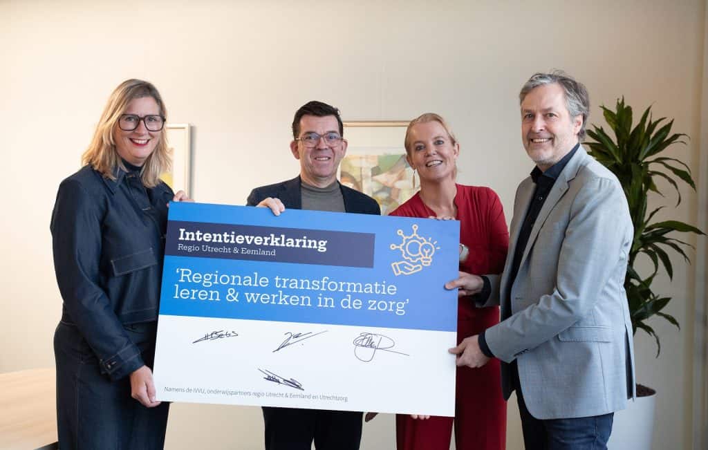 Vier mensen houden een bord vast waarop staat "Intentieverklaring Regio Utrecht & Eemland, 'Regionale transformatie leren & werken in de zorg'". Links staat een vrouw met blond haar en een bril, daarnaast een man met kort bruin haar en een bril, daarnaast een vrouw met een rood pak en blond haar, en helemaal rechts een man met grijs haar en een grijs pak. Ze kijken lachend in de camera.
