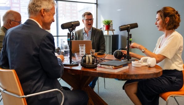 Vier mensen aan een ovale tafel met ieder een microfoon voor zich. Ze nemen een podcast op.
