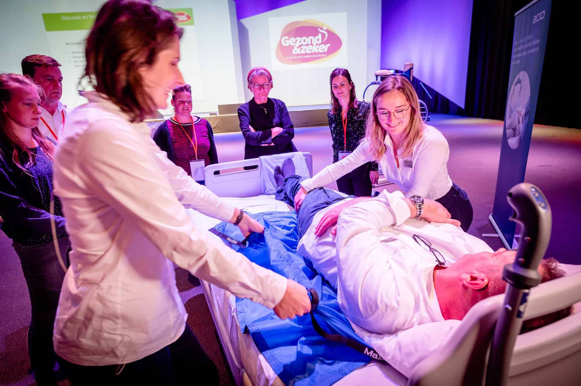 Twee mensen staan om een ziekenhuisbed heen. Er ligt een man in het bed, die door de twee anderen wordt opgetild met een blauw zeil. Verderop staan andere mensen te kijken hoe ze dat doen.