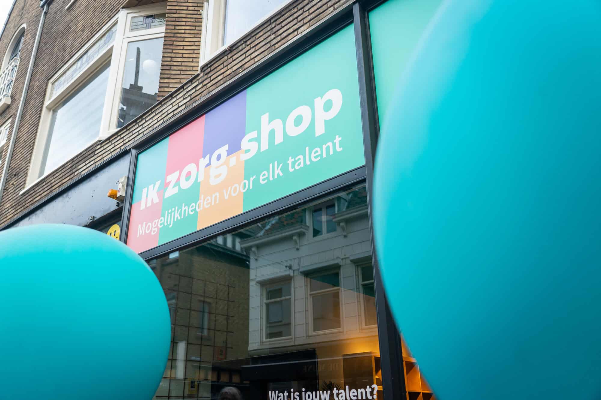 Je ziet de voorgevel van de Ik Zorg Shop, een winkel in Utrecht. Het bord op het gebouw is gekleurd in verschillende blokken, en in witte letters staat er "Ik Zorg Shop, mogelijkheden voor elk talent". Links en rechts op de foto zijn blauwe ballonnen in beeld.