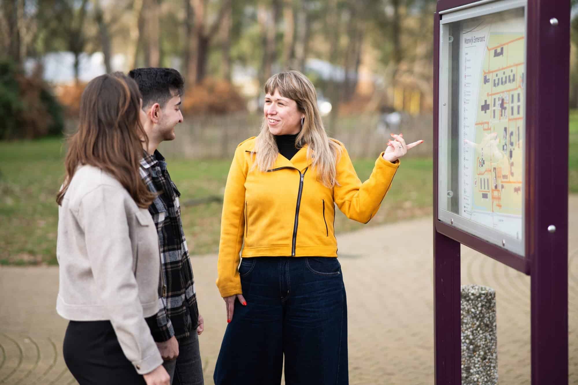 Drie mensen staan buiten, en kijken samen naar een kaart op een bord. Van links naar rechts: een vrouw met lang bruin haar, een man met kort zwart haar, en een vrouw met lang blond haar. De vrouw rechts wijst naar het bord, en kijkt naar de mensen links van haar.
