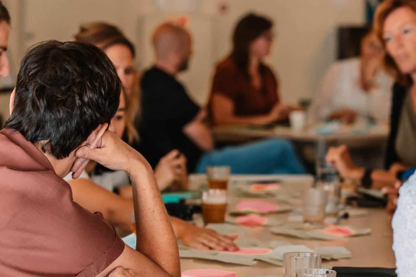 Een groep mensen zit aan tafel. Een man leunt op de tafel, je ziet zijn rug. Er liggen post-its en andere spullen op de tafel.