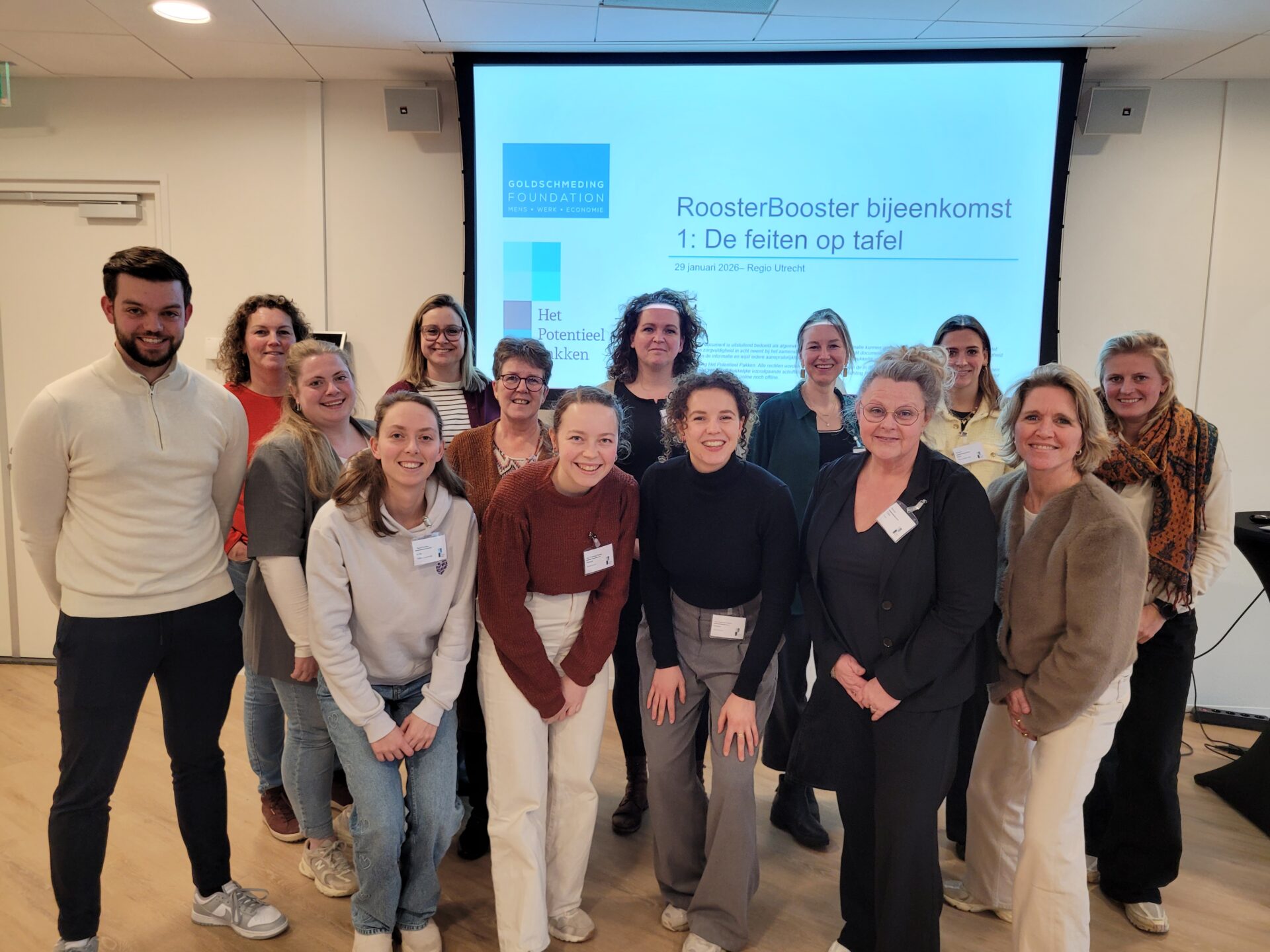 Een groep van ongeveer 20 mensen staat voor een diascherm. Ze kijken lachend naar de camera.