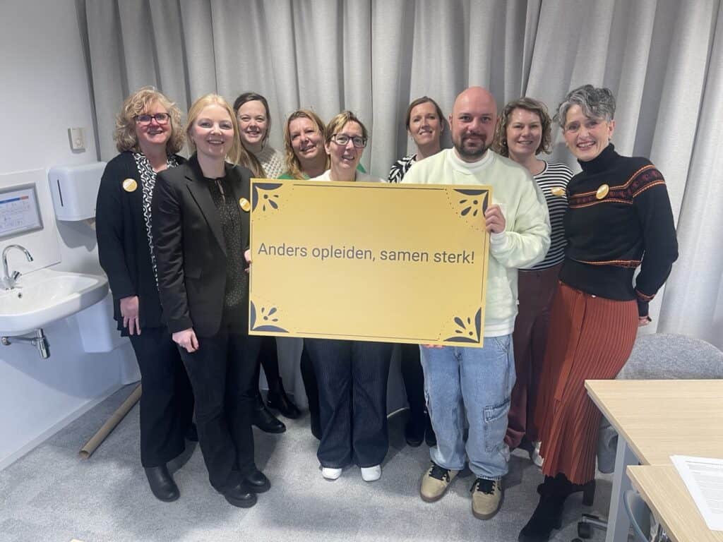 Een groep van 9 mensen staat bij elkaar en kijkt in de camera. Ze houden een bord vast waarop "Anders opleiden, samen sterk!" te lezen is.