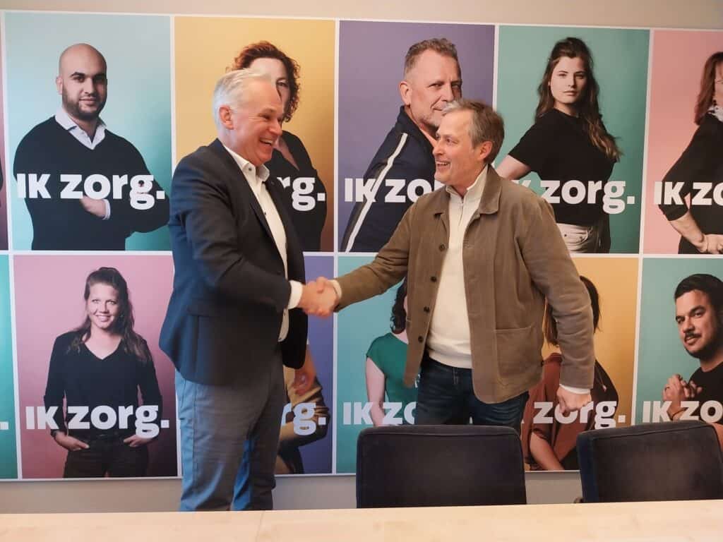 Twee mannen staan voor de Ik Zorg-campagneborden en geven elkaar een hand.