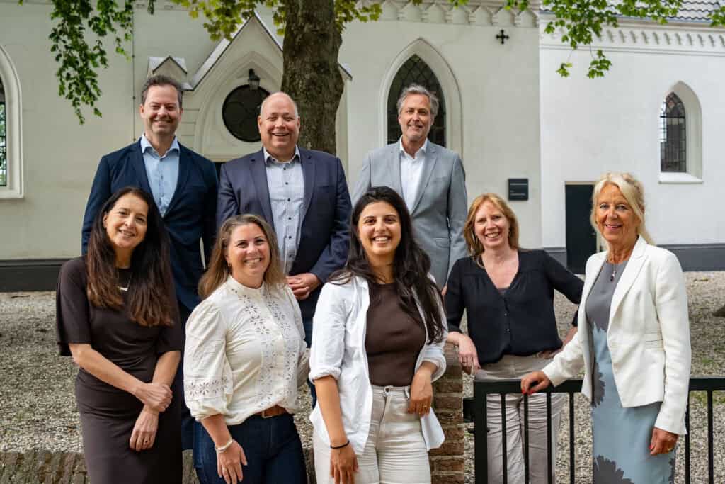 Vijf mensen staan buiten voor een witte kerk, glimlachend en gekleed in zakelijke kleding.